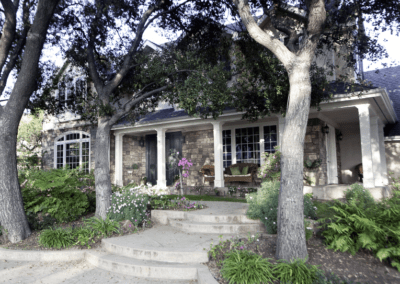 An elegant stone house with a covered porch surrounded by mature trees and landscaped garden.