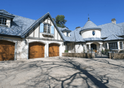 French country-style house with blue-gray shingle roof and three-car garage under clear skies.