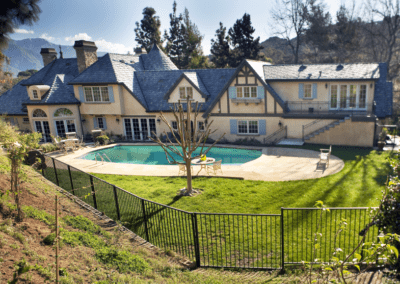 Large two-story house with a french architectural design, featuring a swimming pool and a well-manicured lawn, enclosed by a metal fence.