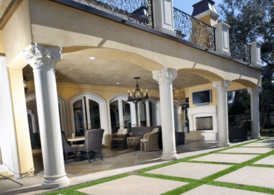 Elegant outdoor patio with classical columns and iron balcony railing, featuring a seating area and a television.